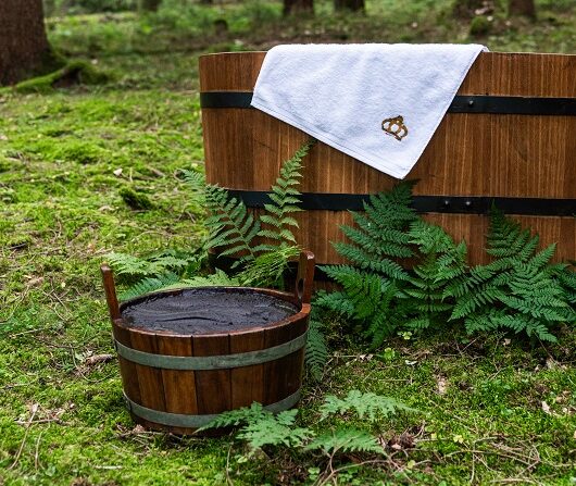 Eine Holzbadewanne steht im Wald mit einem weißen Handtuch und davor steht ein kleiner Holzkübel mit Wasser