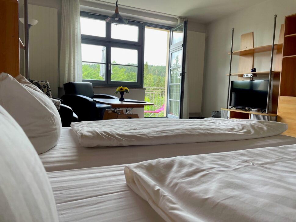 Doppelbettzimmer mit kleinem Tisch, zwei Sessel und einem Fernseher mit Blick ins Grüne