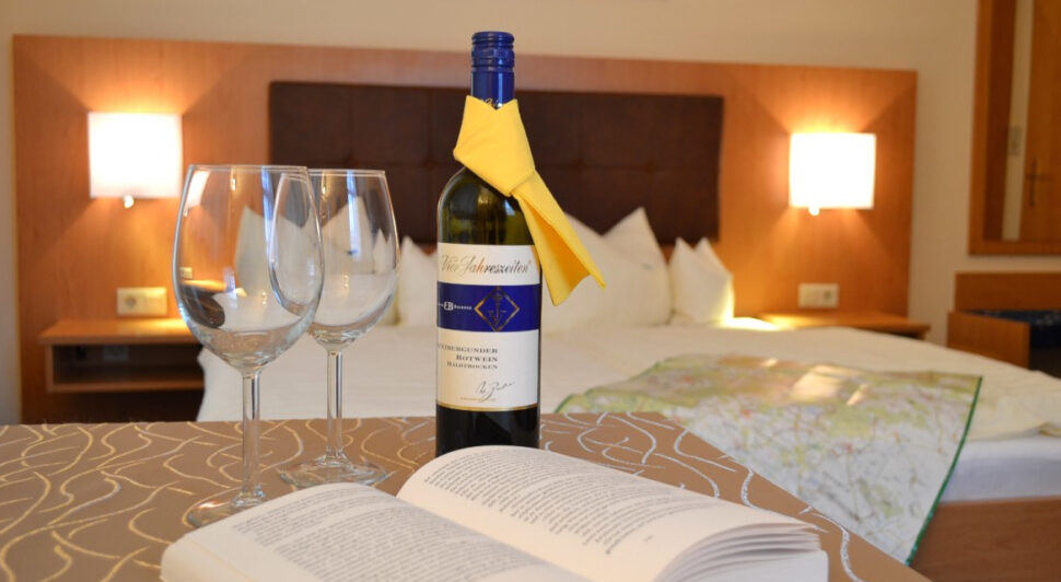 Detailansicht mit einer Flasche Wein undzwei Gläsern mit einem aufgeschlagenem Buch, im Hintergrund ist unscharf ein Doppelbett zu sehen