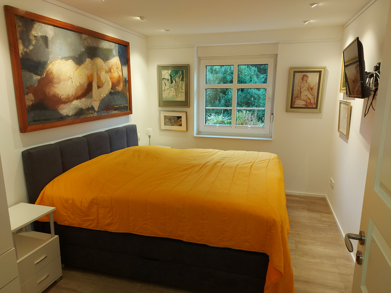 Blick auf ein graues Stoff-Doppelzimmer mit orangener Tagesdecke