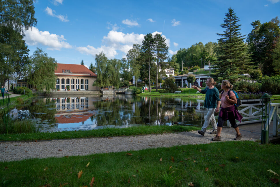 Zwei Personen laufen im Kurpark am Teich entlang, im Hintergrund ist die Festhalle zu sehen