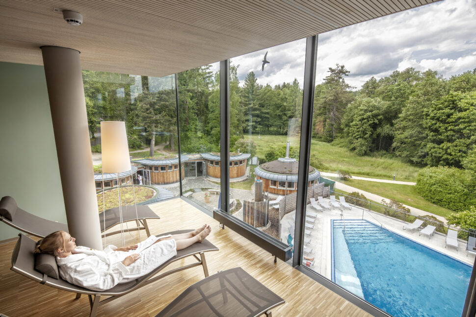 Eine Frau im Bademantel liegt auf einer Liege im Ruheraum des Radon-Therapiezentrums und schaut nach draußen auf die Saunawelt mit Schwimmbecken